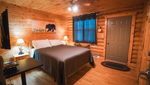 A log cabin bedroom with a bed, two bedside lamps, a ceiling fan, a door, and a window with curtains. - Red Bank Lodge - Red Bank, New Brunswick (Sillikers)