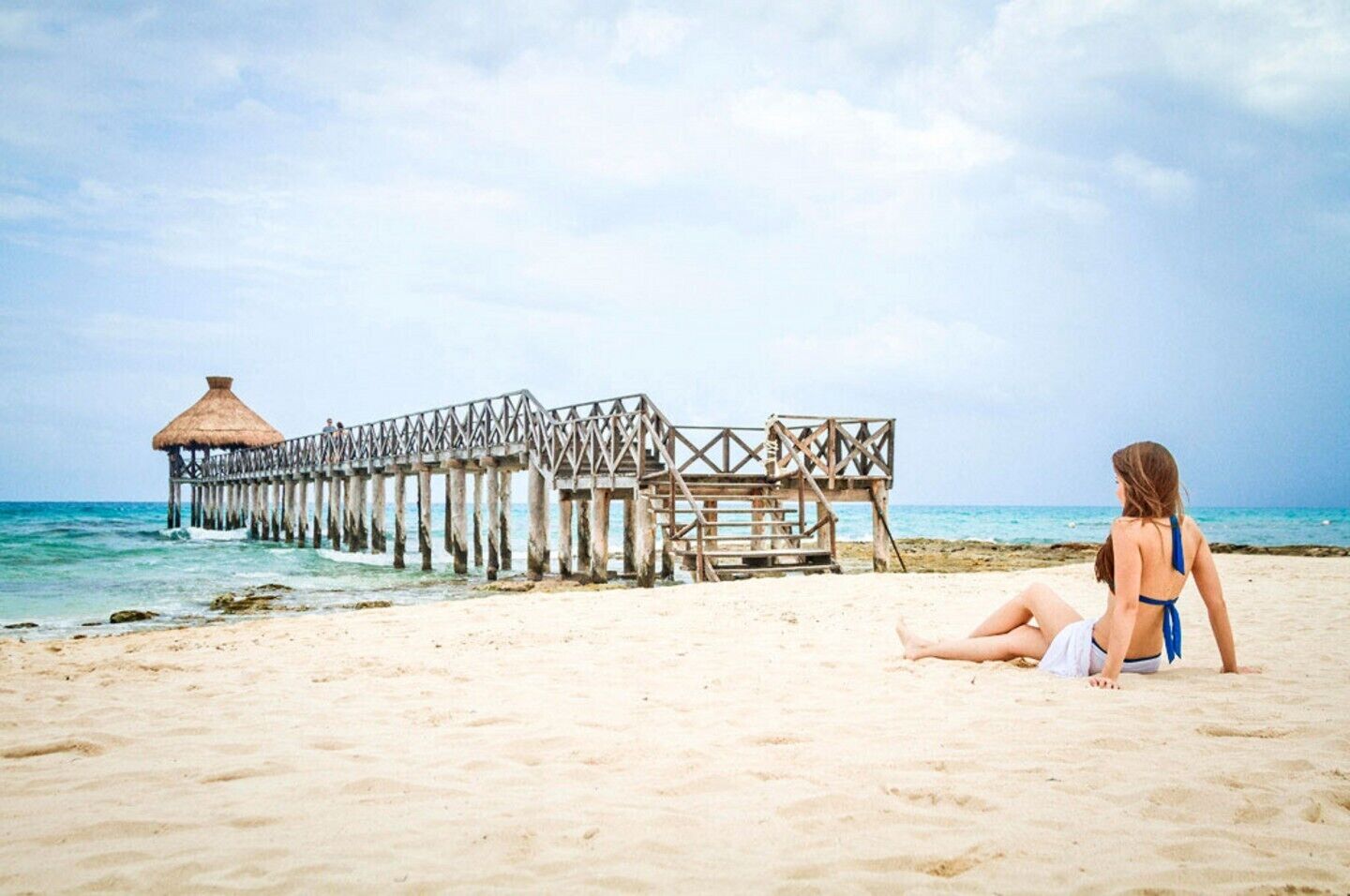 A beach with a wooden pier extending into the sea, a thatched-roof hut at the end, and a woman sitting on the sand.