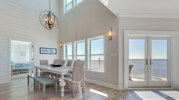 A spacious dining area with a wooden table and wicker chairs, large windows with ocean views, and a chandelier. - Ocean Springs 'Magnolia Beach House' on Pvt Beach! (Ocean Springs)