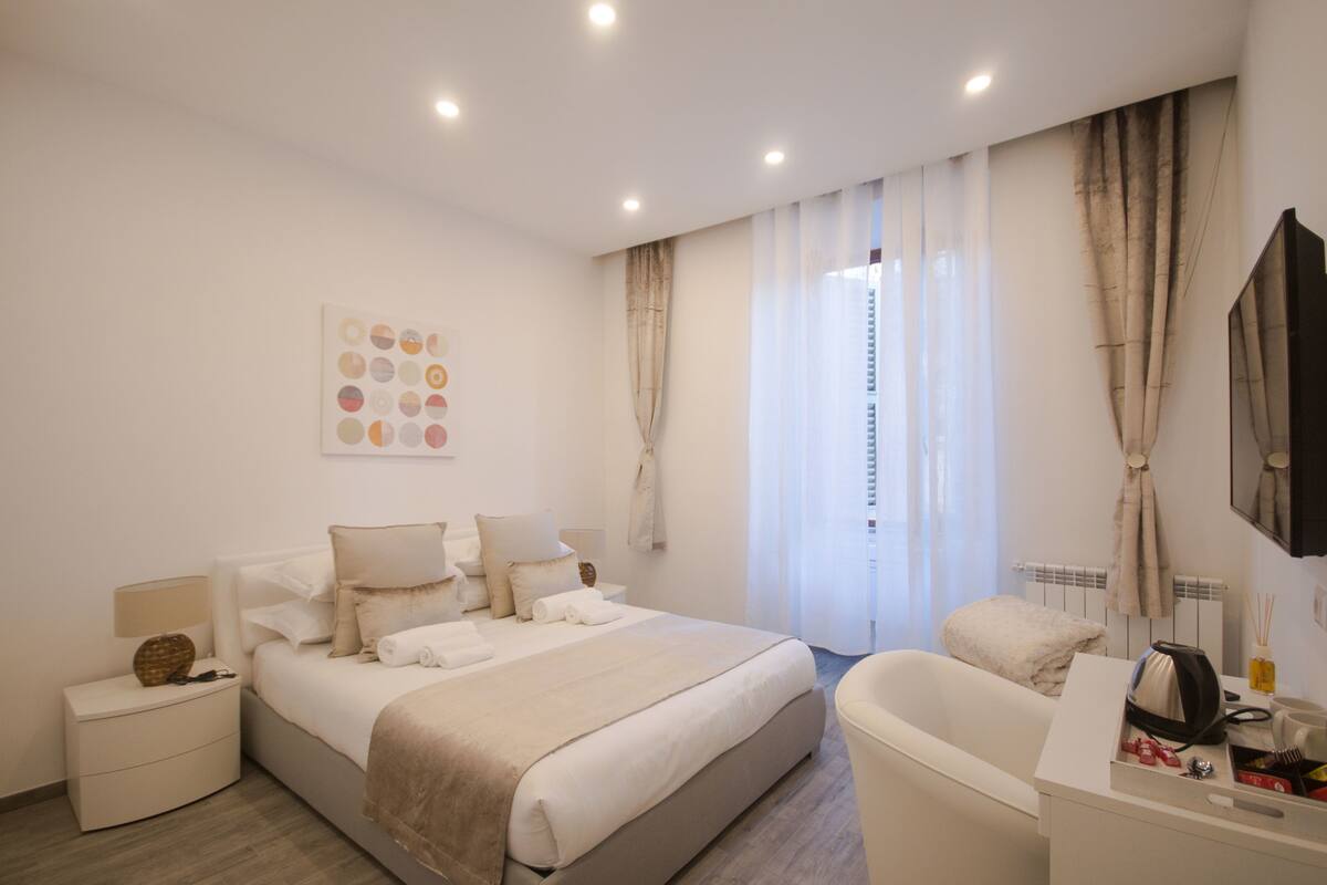 A modern hotel room with a large bed, a bedside table with a lamp, a chair, and a small table with toiletries.