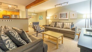 A living room with a sofa, armchair, and coffee table, a kitchen area with appliances, and artwork on the walls.
