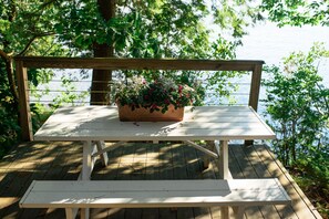 Um deck de madeira com mesa de piquenique e banco, um vaso de flores e vista para a água.