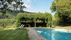 En swimmingpool med en stenhævet sti, der fører til et skyggefuldt siddeområde under en pergola med slyngplanter.