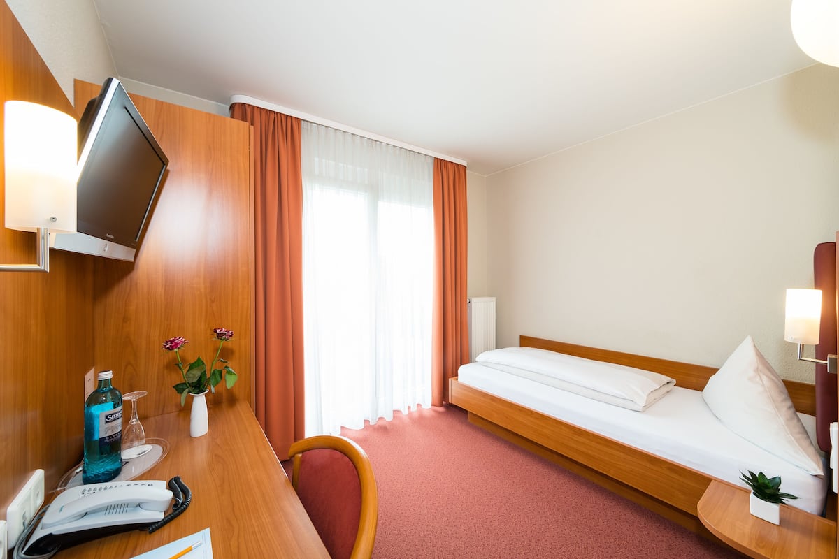 A hotel room with a bed, a desk with a telephone and papers, a TV mounted on the wall, and a window with curtains.
