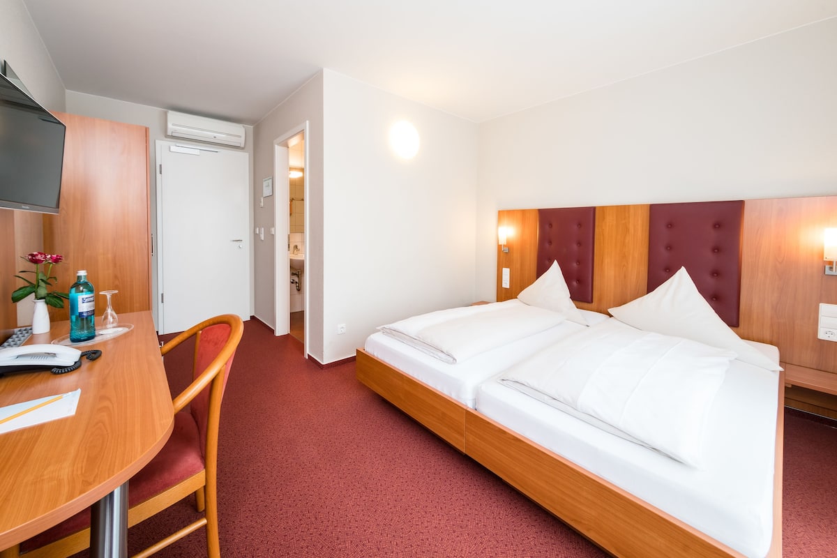 A hotel room with a bed, a desk with a chair, a TV, and a bathroom visible through an open door.
