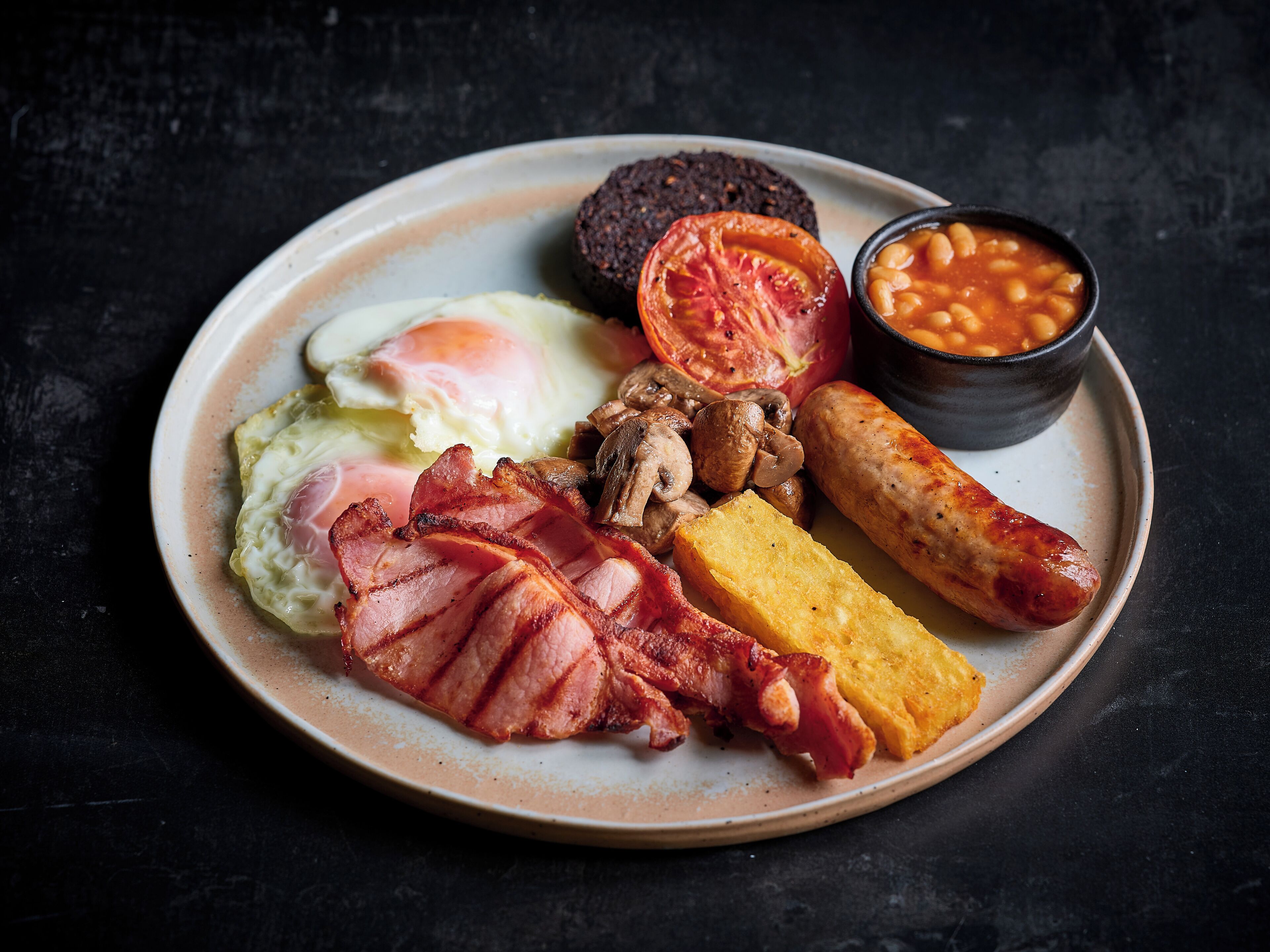 Un plato de comida para el desayuno con huevos, tocino, salchichas, champiñones, tostadas, frijoles horneados y tomate.
