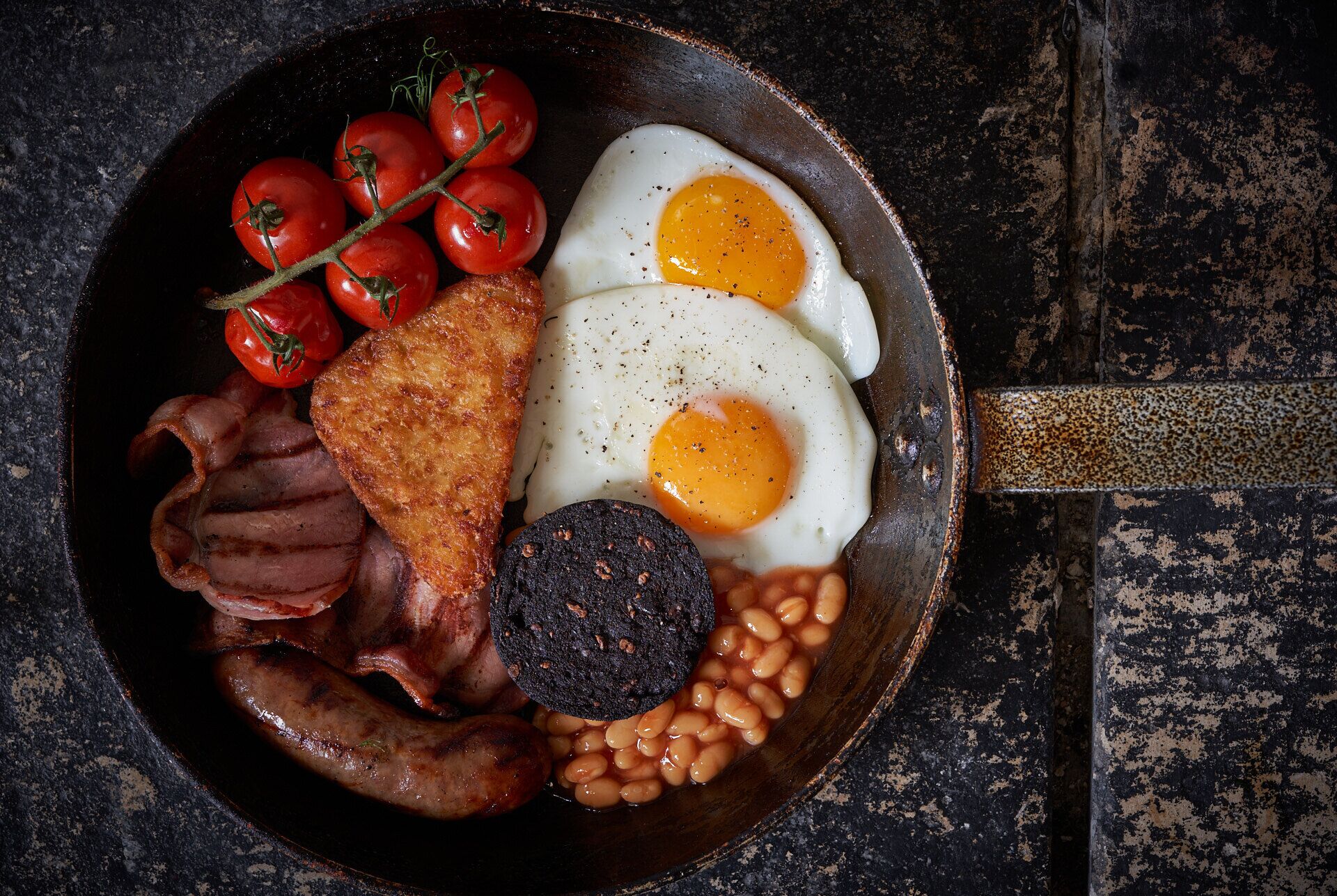 Un plato de desayuno con huevos, tocino, salchichas, tomates, morcilla y frijoles horneados.