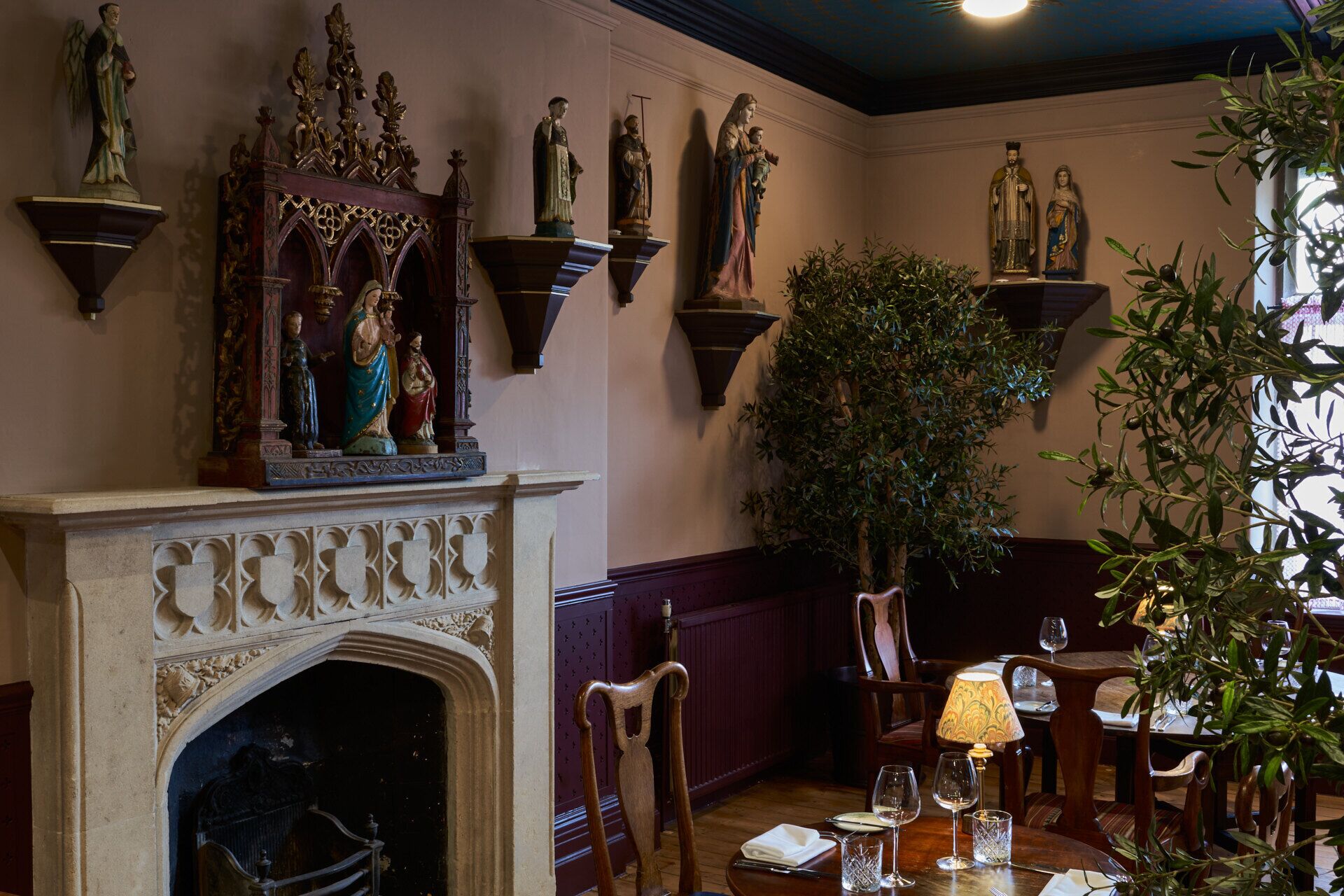 A room with a fireplace, wooden chairs, a dining table with wine glasses, and wall-mounted statues.
