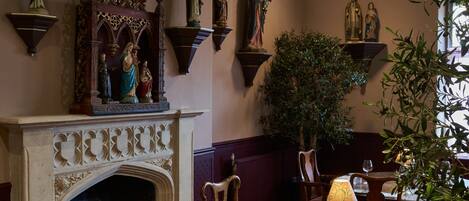 A room with a fireplace, wooden chairs, a dining table with wine glasses, and wall-mounted statues.