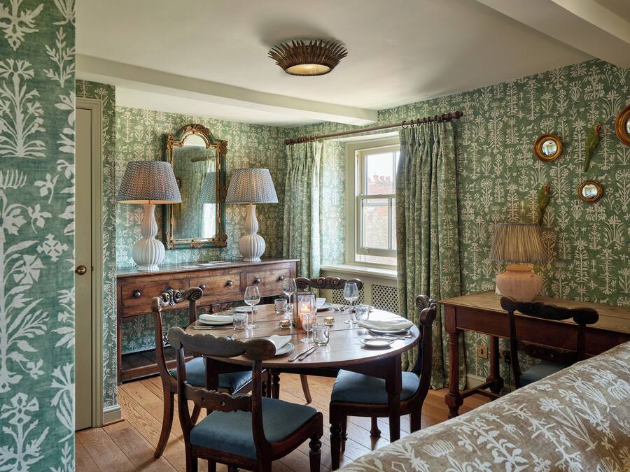 A dining room with a wooden table set for four, surrounded by upholstered chairs. A wooden dresser with a mirror and lamps is against the wall.