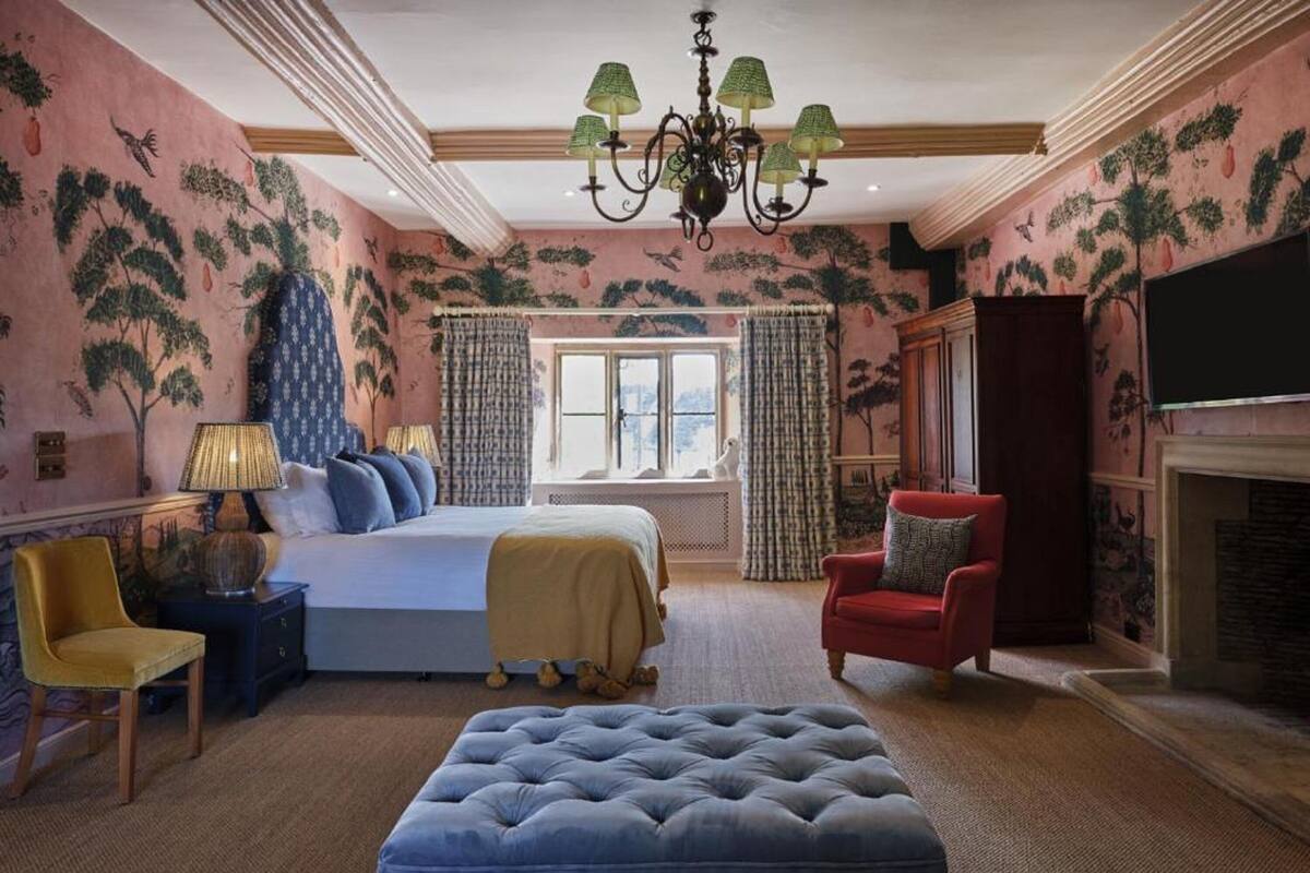 A bedroom with a bed, a red armchair, a blue ottoman, a chandelier, and a fireplace.