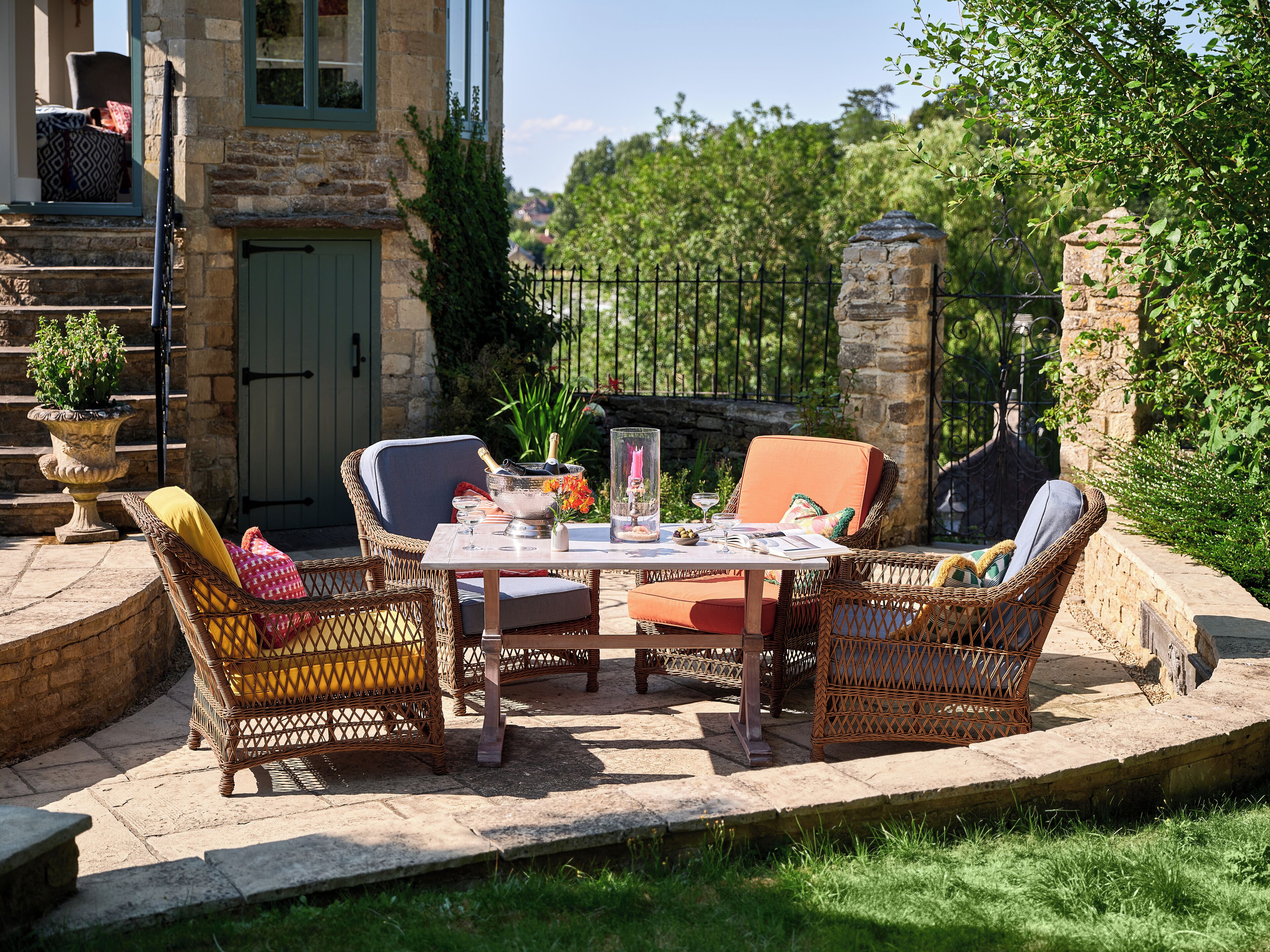 An outdoor patio with wicker furniture, a table set with glasses and a bottle, and a stone wall in the background.