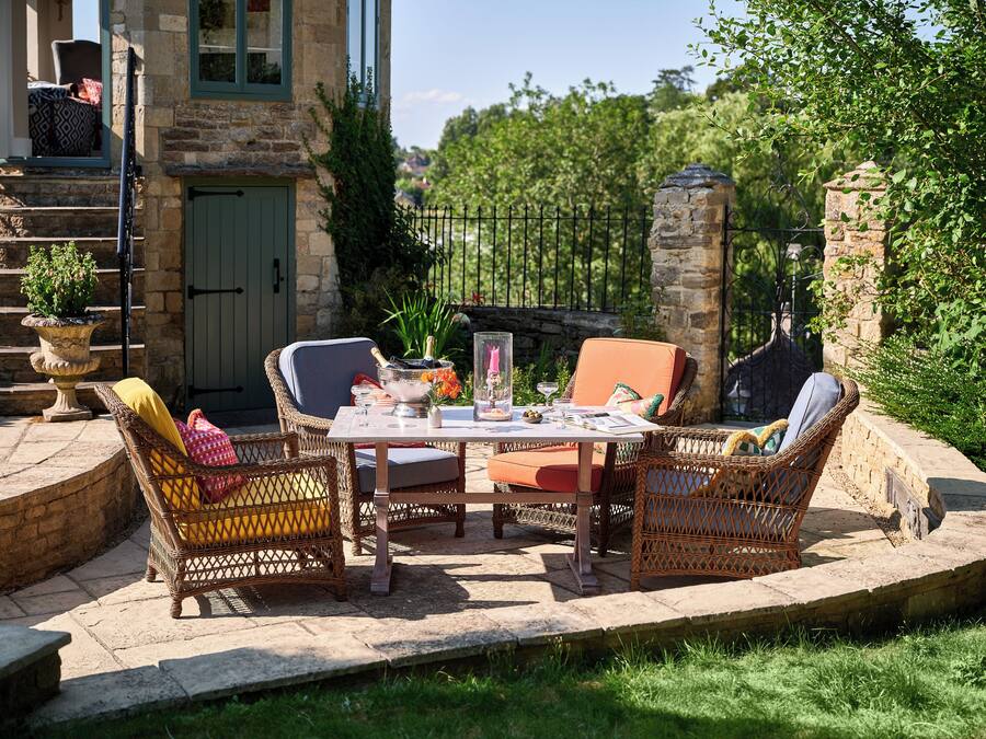 An outdoor patio with wicker furniture, a table set with glasses and a bottle, and a stone wall in the background.