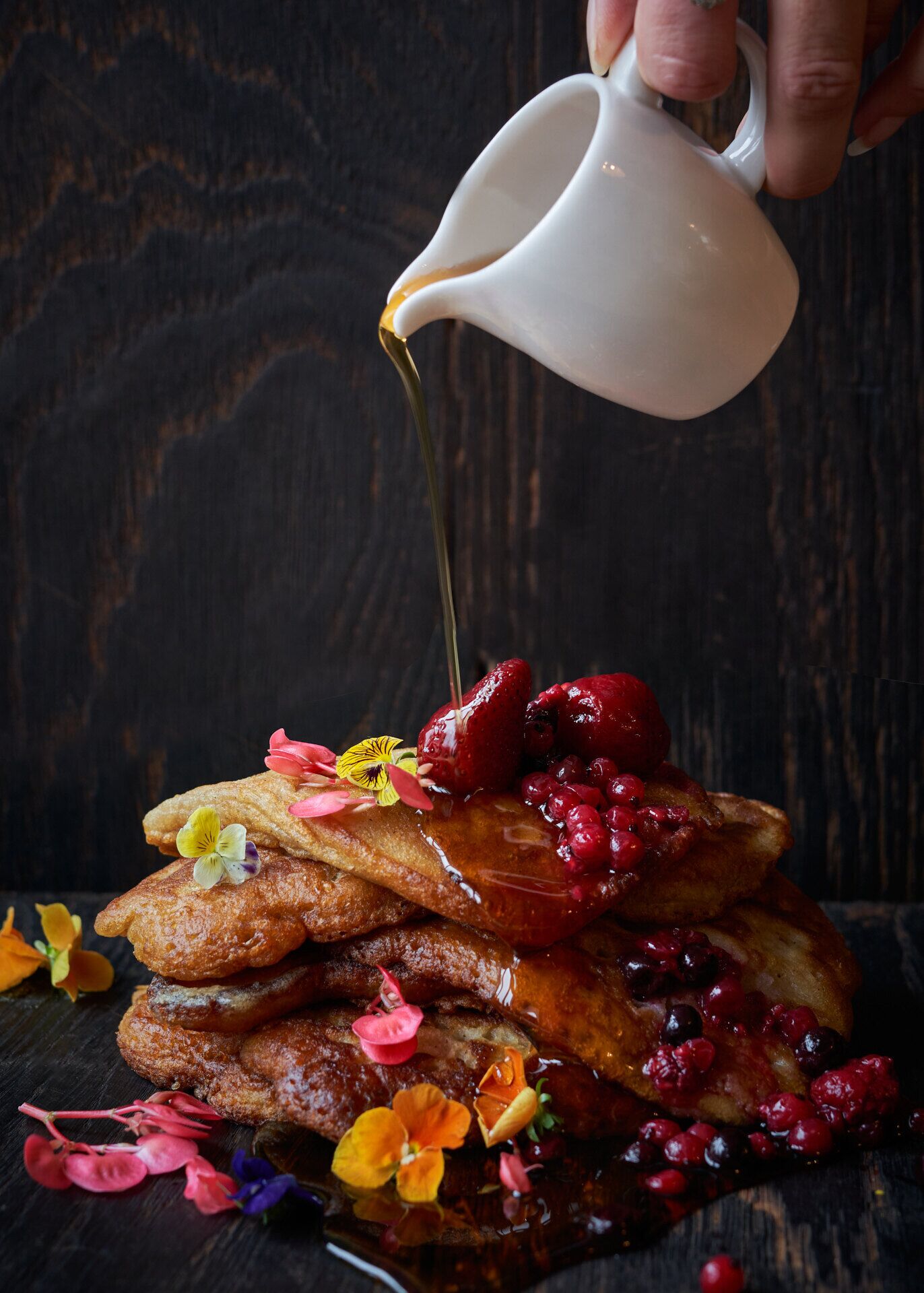 A stack of pancakes with syrup being poured over them, garnished with berries and flowers.
