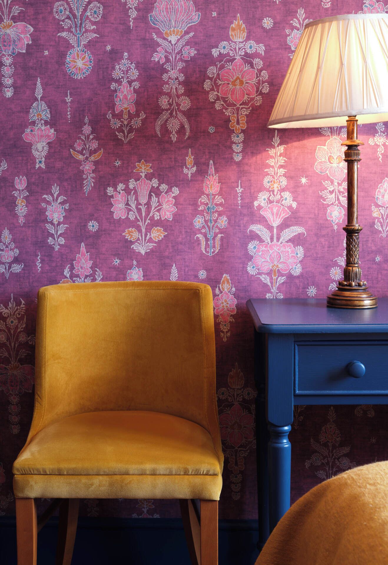 A room with a velvet armchair, a blue nightstand with a lamp, and a floral-patterned wallpaper.