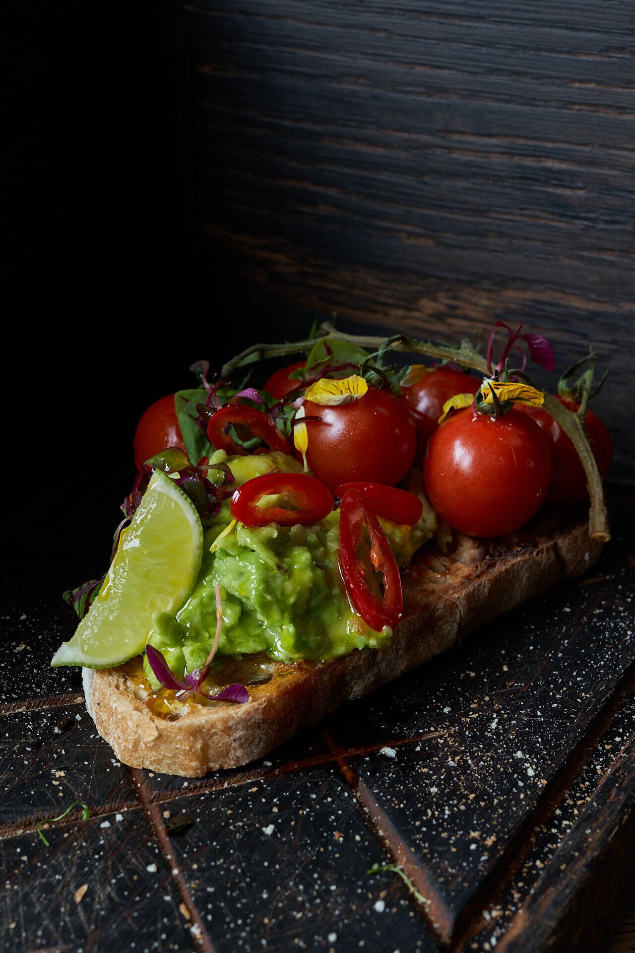 Un sándwich con aguacate, tomates y lima sobre una rebanada de pan, sobre una superficie de madera oscura.