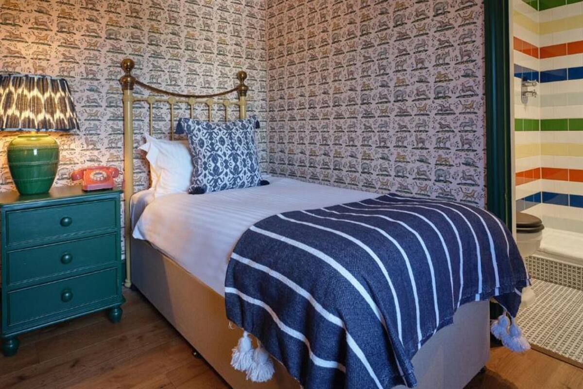 A bedroom with a brick-patterned wall, a bed with a striped blanket, a green dresser, a lamp, and a bathroom with colorful tiles.