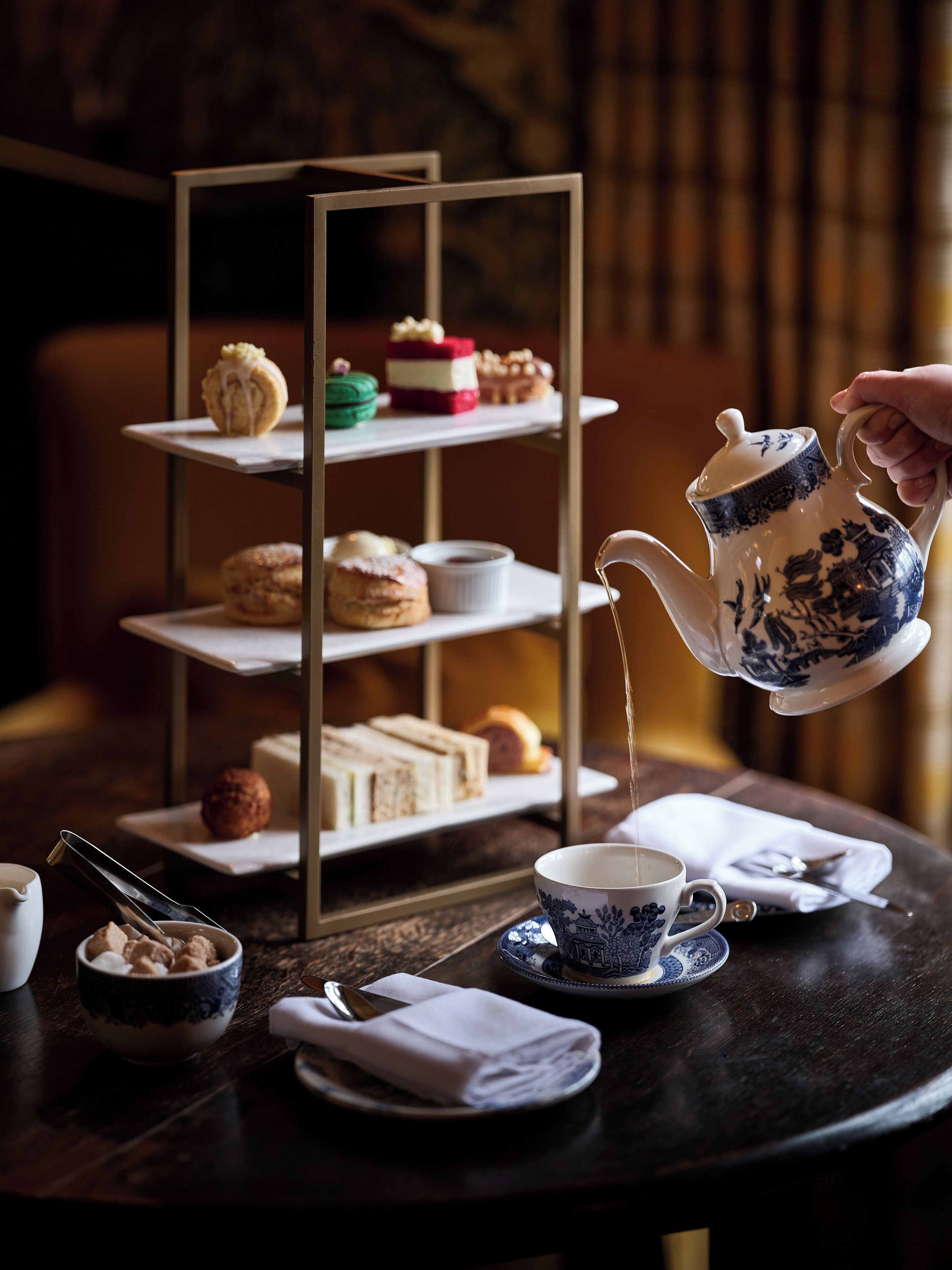 Un servicio de té con una tetera sirviendo en una taza, un pastelero de postres en varios niveles y varias tazas y platillos para té sobre una mesa.