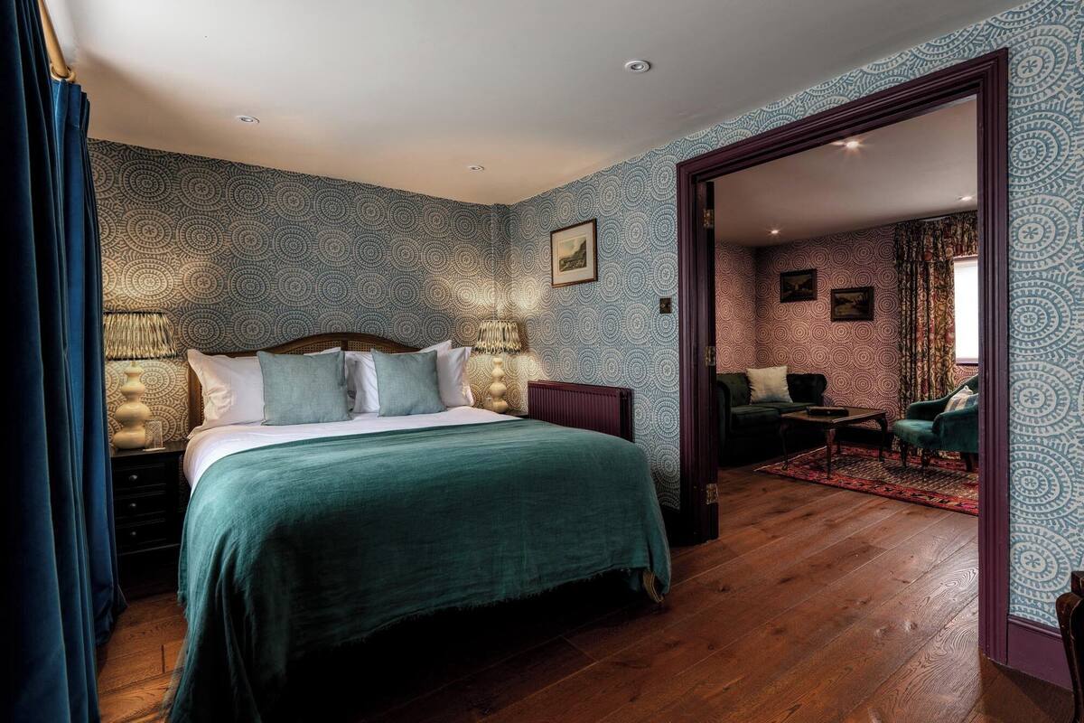A bedroom with a large bed, a wooden floor, patterned wallpaper, and a view into a living area with a sofa and a coffee table.