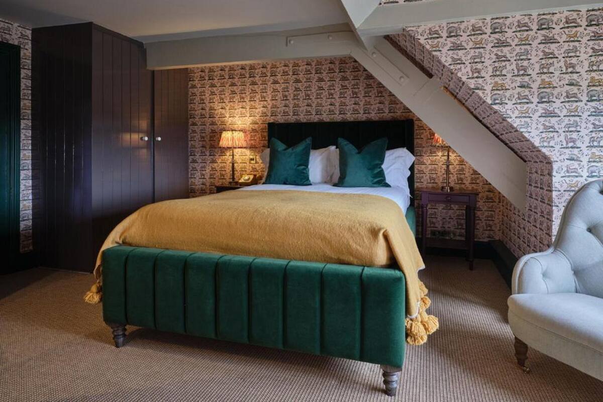 A bedroom with a green upholstered bed, a dark wardrobe, a nightstand with a lamp, and a patterned wallpaper.