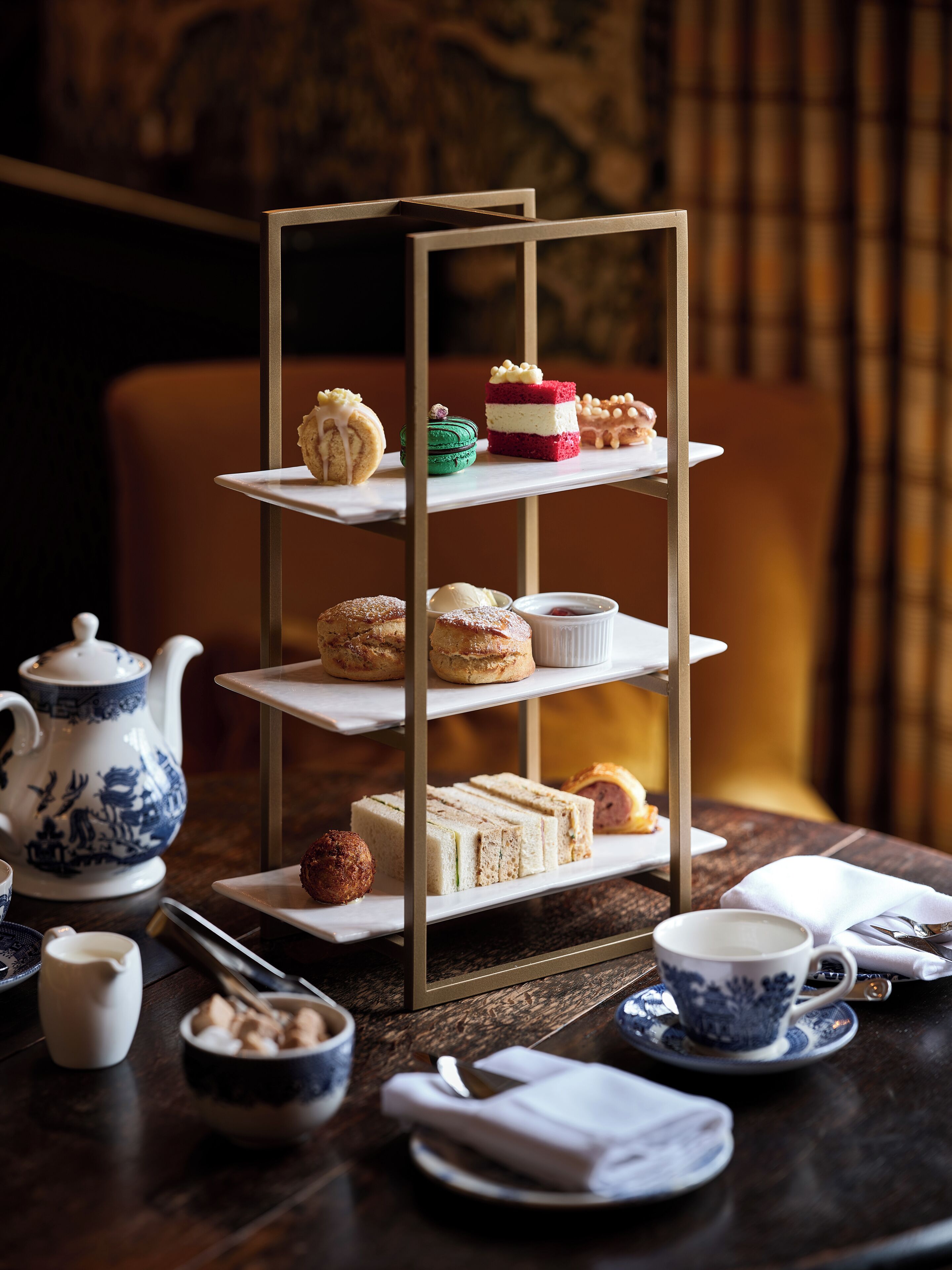A three-tiered dessert stand with various pastries and cakes, a teapot, cups, and a napkin on a wooden table.