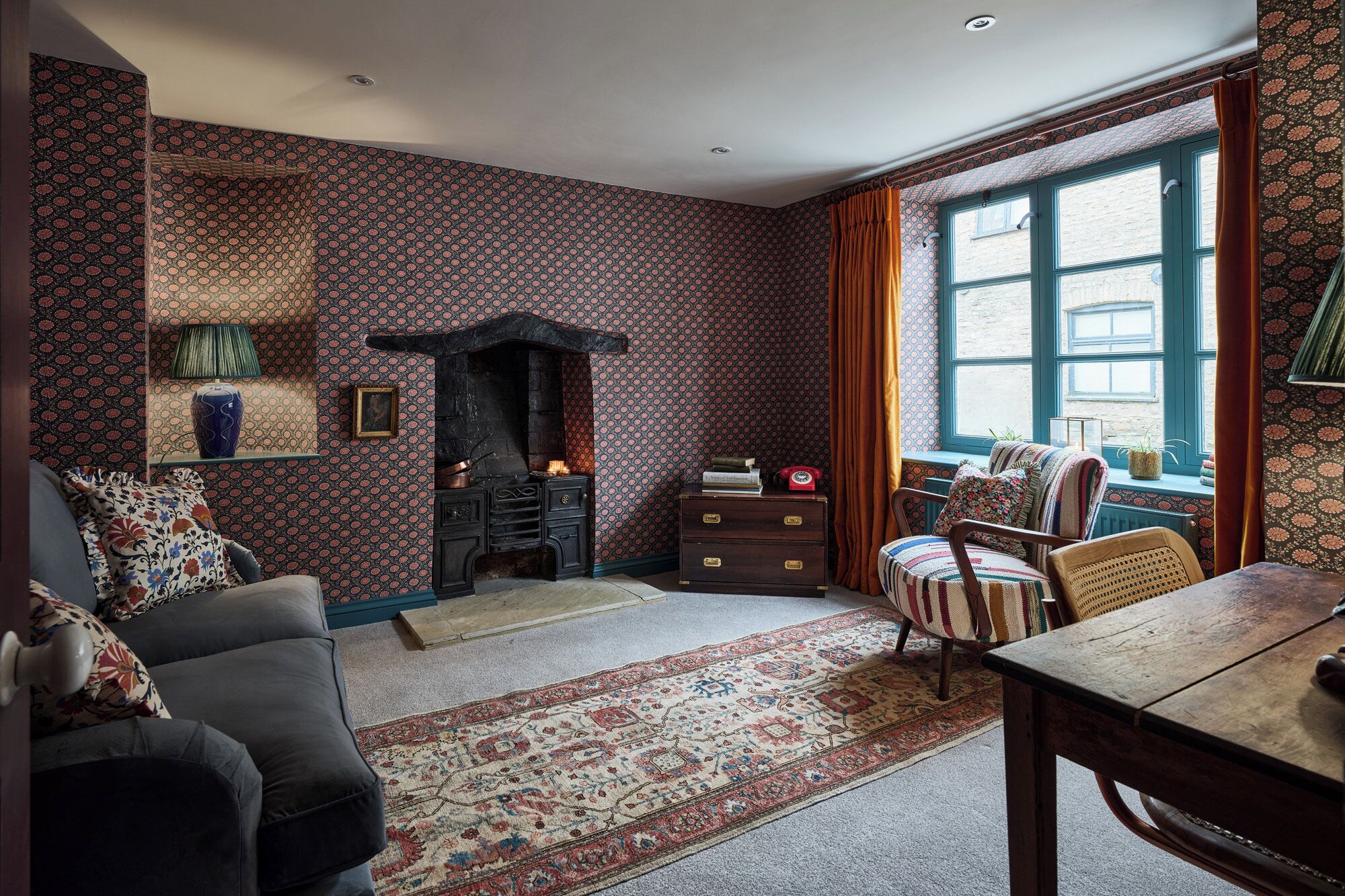 A traditional room with patterned wallpaper, a fireplace, a sofa, a chair, and a window with curtains.