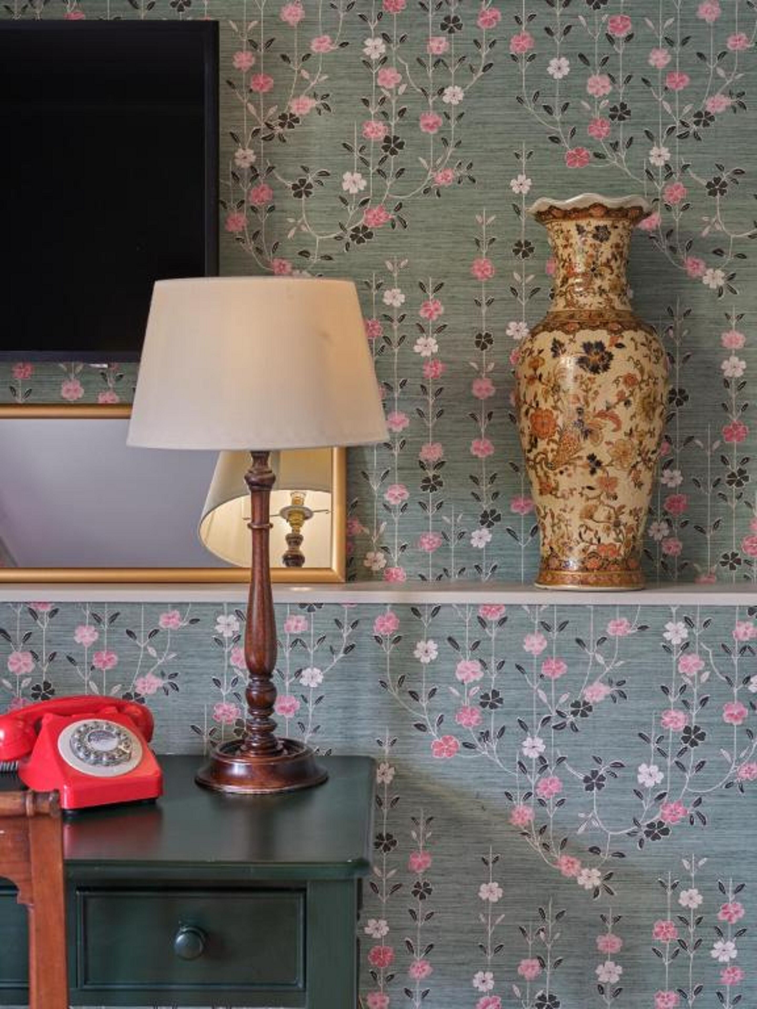 Una habitación con papel tapiz floral, una lámpara, un jarrón y un teléfono rotatorio rojo sobre un aparador.