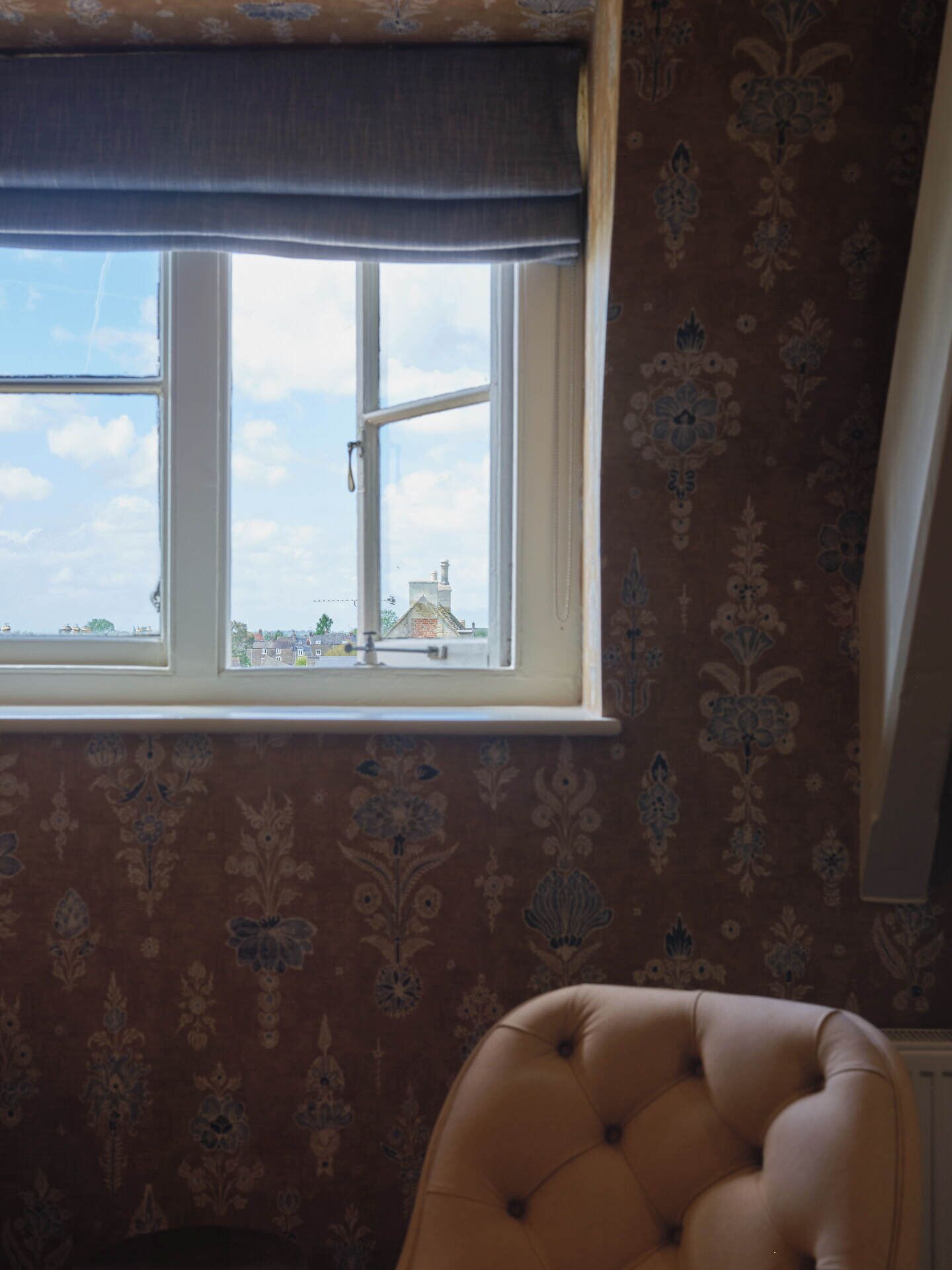 A room with a window, patterned wallpaper, and a tufted chair.