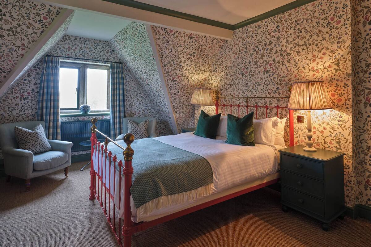 A bedroom with a sloped ceiling, floral wallpaper, a bed with green and white bedding, a nightstand, a chair, and a window with curtains.
