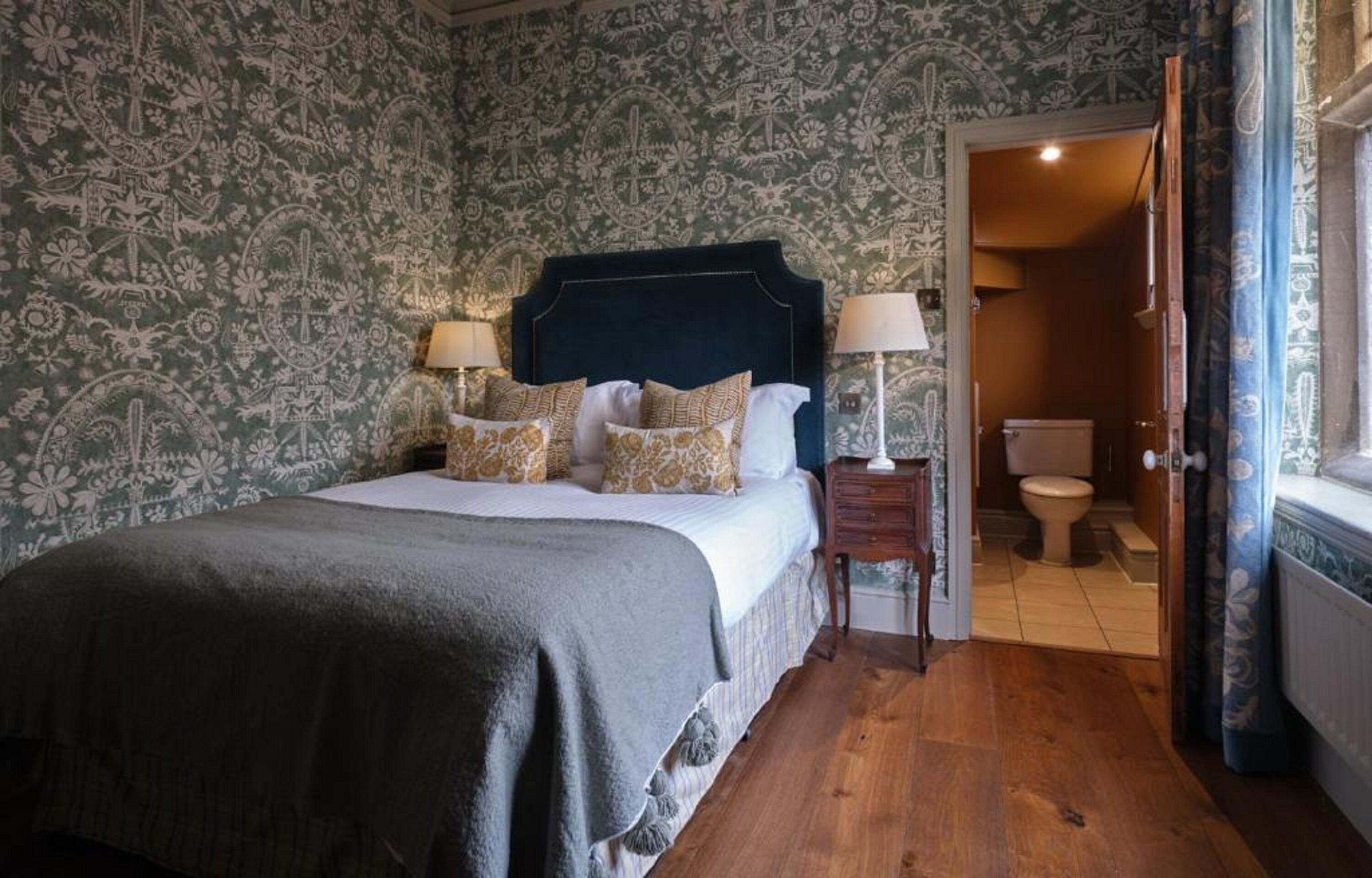 A bedroom with a large bed, two bedside tables with lamps, a wooden floor, and a patterned wallpaper.