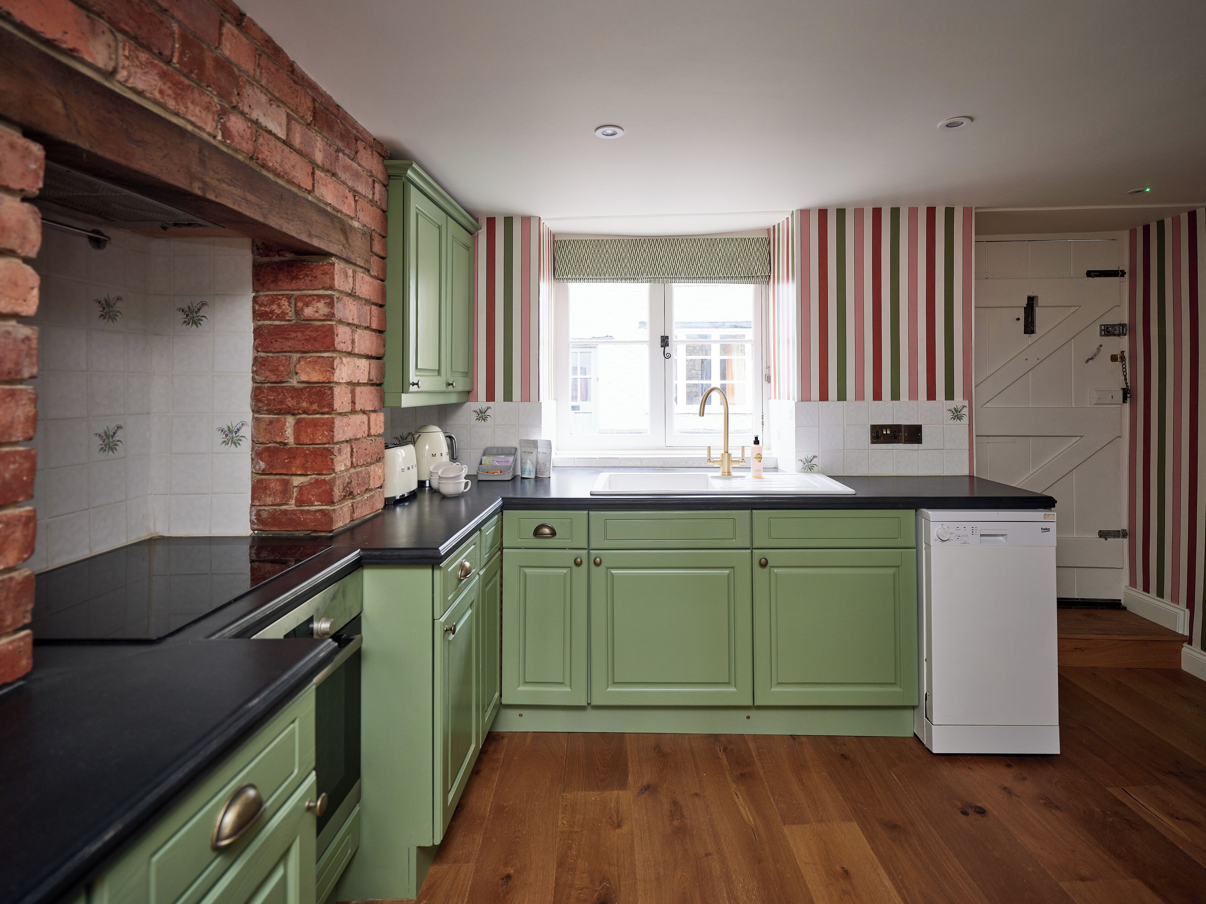 Une cuisine avec des meubles de couleur verte, un mur en briques, un plancher en bois, un lave-vaisselle blanc et un papier peint à rayures.