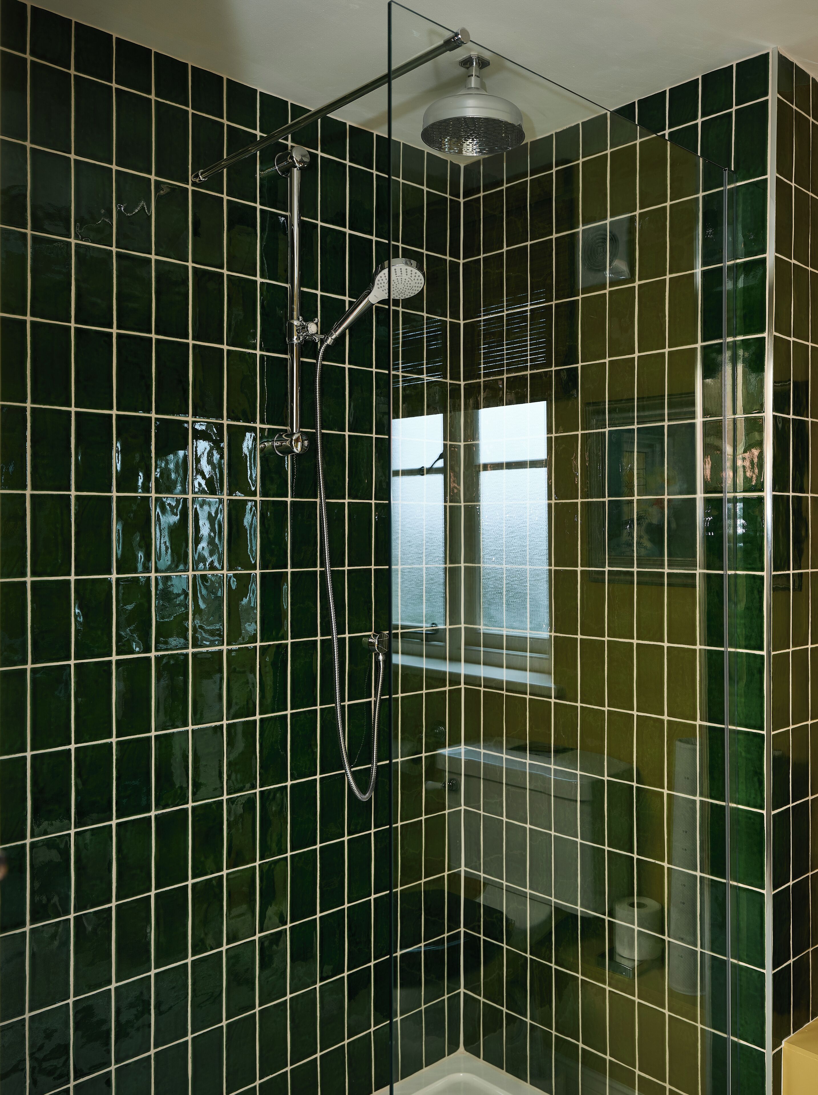 Une salle de bain avec des carreaux vert foncé, une douche en verre et un porte-rouleau de papier toilette visible.