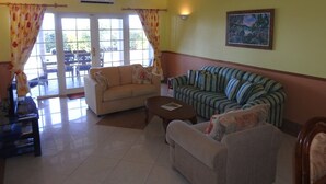 A living room with a beige sofa, a striped sofa, a coffee table, and a ceiling fan. There is a window with curtains, a painting on the wall, and an air conditioning unit.