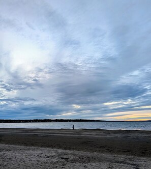 A beach with a person walking, a distant shoreline, and a cloudy sky.