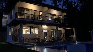 Une maison moderne à deux étages, dotée d’une piscine, de grandes fenêtres et d’un espace salon extérieur.