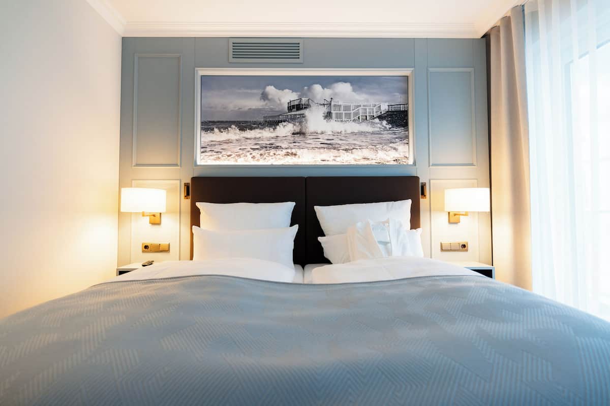 Habitación de hotel con una cama grande, dos lámparas de noche, un cabecero y un televisor empotrado en la pared mostrando una escena costera.