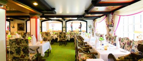 A dining area with floral-patterned chairs and tables, green carpet, and a high ceiling with exposed beams and floral wallpaper.