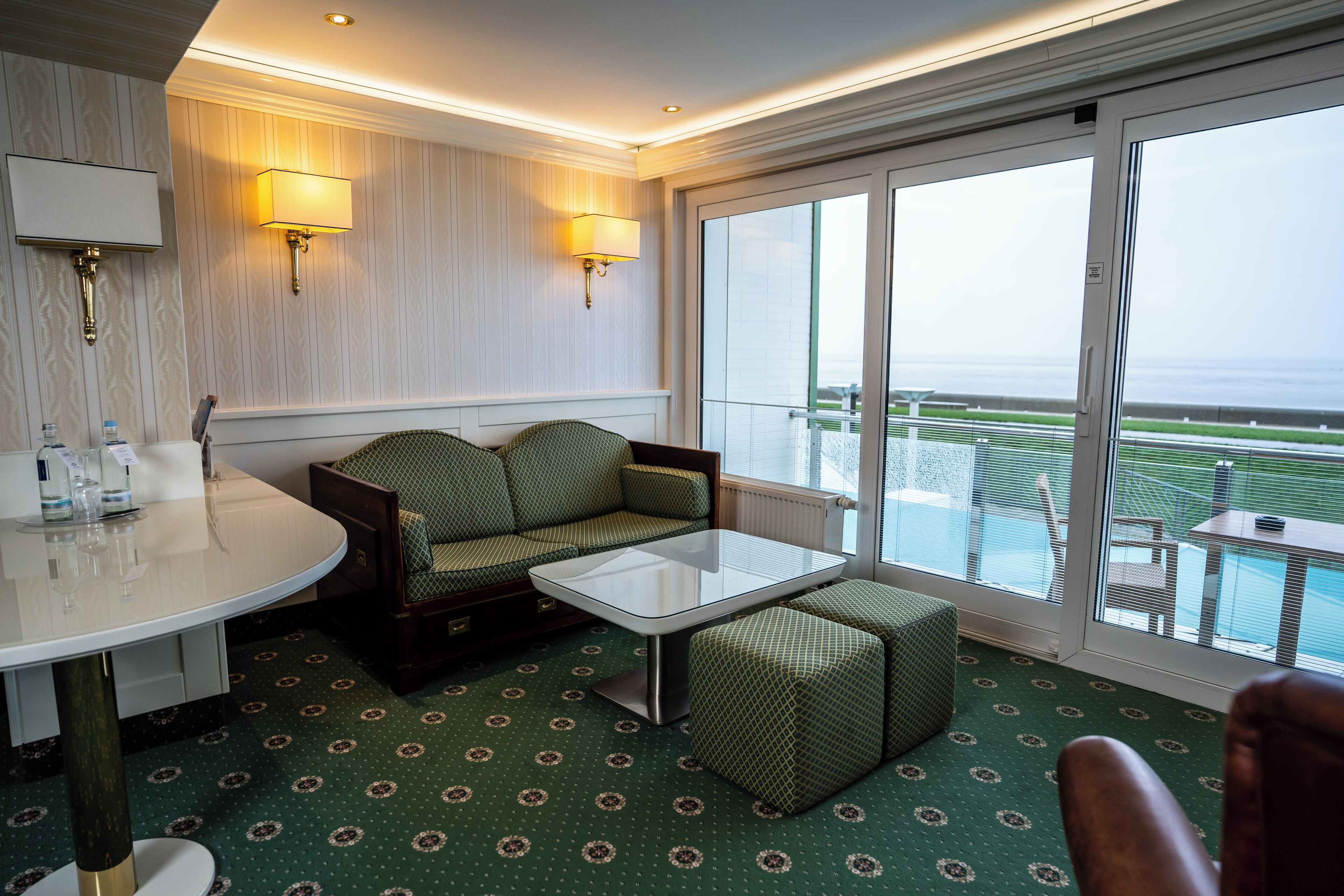 Um quarto com carpete verde estampado, uma mesa redonda com tampo de vidro, um sofá verde, um banquinho combinando e duas luminárias fixadas na parede.