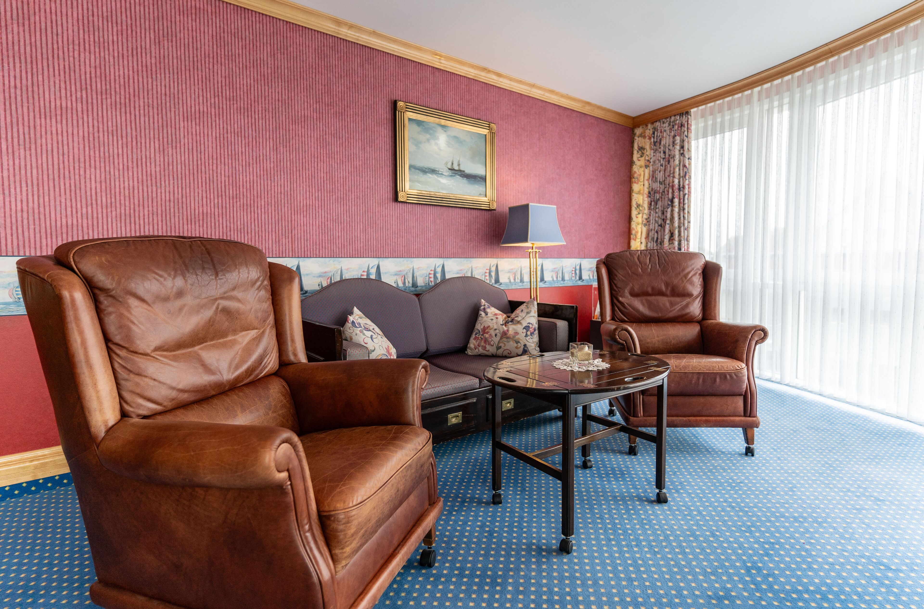 Hotellihuone, jossa on kaksi nahkaista nojatuolia, sohva, pieni pöytä lampun kanssa ja suuri ikkuna, jossa on läpikuultavat verhot.
