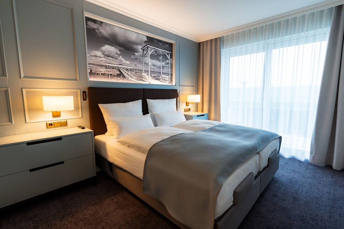 Habitación de hotel con una cama grande, dos mesitas de noche con lámparas, un cabecero, un cuadro enmarcado en la pared y una ventana con cortinas.