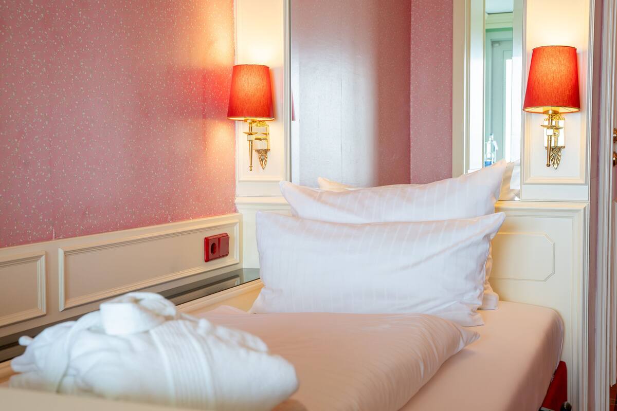 Una habitación de hotel con una cama bien hecha, dos lámparas fijadas a la pared y un teléfono rojo en la mesita de noche.
