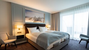 A hotel room with a large bed, two bedside tables with lamps, a chair, a large window with curtains, and a painting above the bed. - Hotel Strandperle (Cuxhaven)