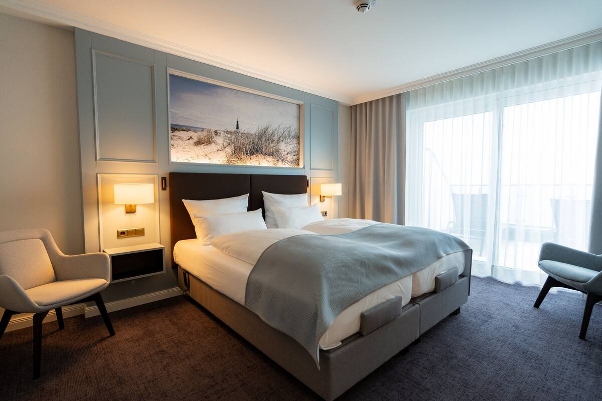 Habitación de hotel con una cama grande, dos mesitas de noche con lámparas, una silla, un ventanal con cortinas y un cuadro sobre la cama.
