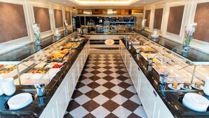 A buffet area with a checkered floor, glass display cases with food, and a bar in the background. - Hotel Strandperle (Cuxhaven)