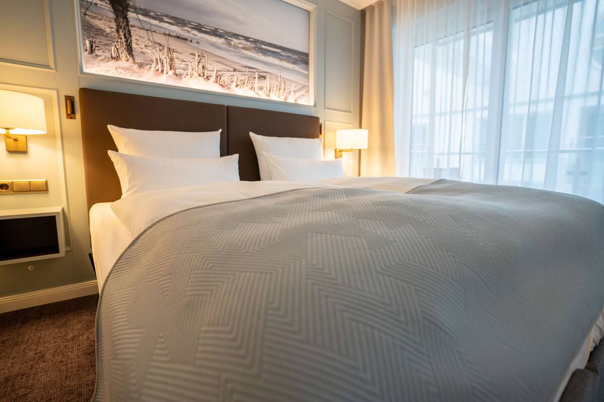 Habitación de hotel con una cama grande, dos lámparas de noche, un cabecero con una pintura de paisaje y una ventana con cortinas traslúcidas.