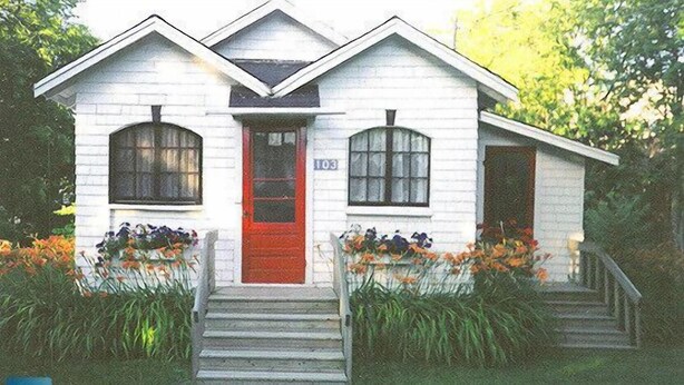 A white house with a red door, numbered 103, and a front porch with steps leading to the entrance.