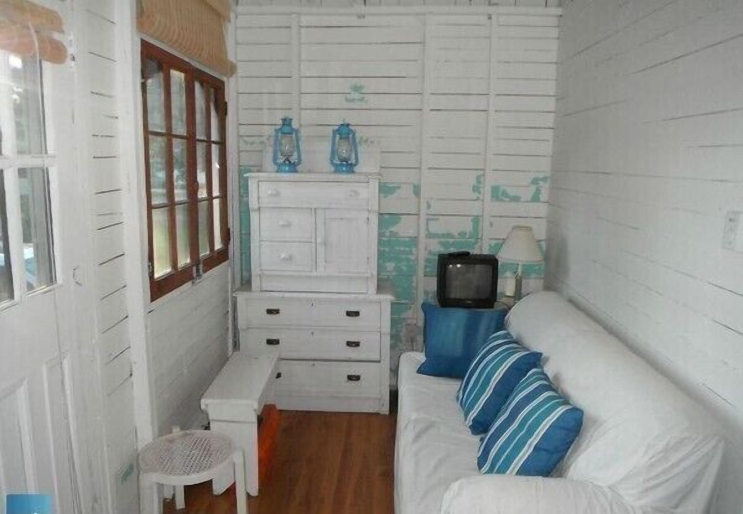 A small, compact room with white walls, a wooden floor, a white sofa with blue stripes, a small white table, a white dresser, and a television on a blue stand.