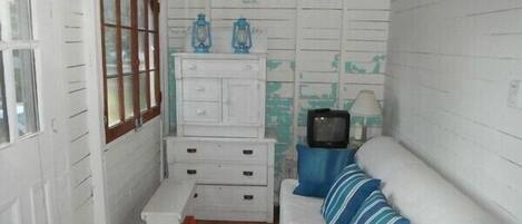 A small, compact room with white walls, a wooden floor, a white sofa with blue stripes, a small white table, a white dresser, and a television on a blue stand.