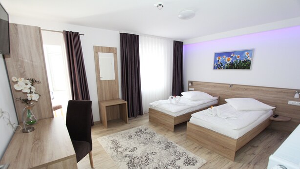 A hotel room with two beds, a desk, a chair, a large window with curtains, a painting on the wall, and a wooden floor. - Pension Villa Colosseo (Meiningen)