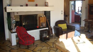Ein gemütliches Wohnzimmer mit Kamin, einem rot karierten Sessel, einem braunen Ledersessel mit gelbem Kissen und einem steinbelagten Boden.